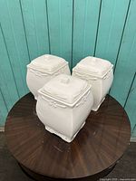 Top view of three white ceramic square canisters placed on a dark wood round table against turquoise wooden paneling.