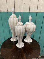 Overall view of three white ceramic pointed lid jars on a round wooden table against teal and white wood plank wall.