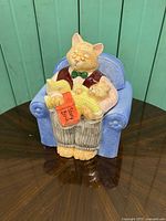 Front view of the hand painted porcelain cat grandpa cookie jar seated in blue armchair holding kitten and story book.