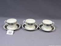 Three sets of Wedgwood tea cups and saucers arranged in a row, all white with black geometric and gold floral patterns.