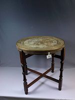 Full view of antique round brass gaming table on dark wood folding stand with cross stretchers and turned legs.