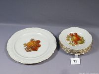 Photo showing the larger plate and the stack of eight smaller plates with fruit and nut designs and scalloped edges.