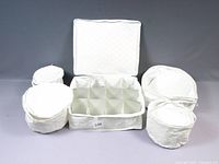 Photo shows one large rectangular white fabric zip up china storage case with twelve compartments inside, and five smaller round white fabric zip up storage cases for smaller items or delicate china.