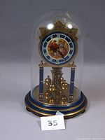 Full view of the anniversary clock inside a glass dome showing blue and gold tones, pendulum balls, and vintage dial illustration.
