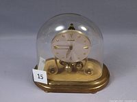 Front view of the oval mechanical anniversary clock under glass dome with gold-tone base and four rotating spheres pendulum.