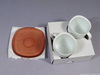 Two white square-shaped ceramic cappuccino mugs on white cardboard holder positioned next to two translucent amber square saucers on white paper.