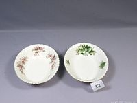 Top view of two oval serving bowls showing floral patterns Lavender Rose on left and Trillium on right, with scalloped edges and gold rims, placed side by side on gray surface.
