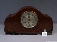 Wooden mantel clock with curved top and brown finish, showing the front and base with carved details.