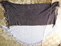 Both shawls laid flat showing black and silver rectangular shawl on top, white semi-circular lace-patterned shawl underneath, fringes visible on edges