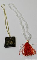 Photo of two long necklaces laid out, showing the floral enamel pendant and clear beads with tassel
