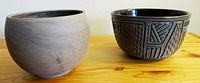 Photo showing two pottery bowls side by side: left is a smooth, light gray Lune Sphere basalt bowl; right is a black-glazed bowl with textured geometric patterns