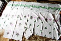 Photo showing two tablecloths and the matching mat, folded napkins in front. The large clover design tablecloth extends across the top with the Lily of the Valley tablecloth partially overlaid below. Napkins are stacked in the front.