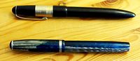 Photo showing two vintage fountain pens placed horizontally on wooden surface, the top pen is black with silver band on cap and the lower pen is blue marbled with silver bands on cap and barrel.