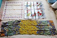 Three floral pattern scarves laid out on a carpet: a long multicolor scarf with Monet iris design at bottom, a silvery scarf with red flowers in the middle right, and a small cream scarf with pastel flowers at top left.
