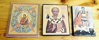 Three small religious icon images arranged on wooden floor, showing Virgin Mary, a saint bishop, and an armored saint with spear.