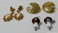Three pairs of earrings shown on white background: gold-tone pierced earrings with faux pearls and rhinestones, gold-tone swirl design clip-ons, and red and white mushroom-shaped pierced earrings.