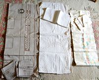Three sets of tablecloths with matching napkins laid out side by side on carpet floor. Left set: beige damask tablecloth with geometric and leaf designs and six matching napkins. Center set: white oval tablecloth with cutwork and embroidery, napkins partially folded on top. Right set: satiny damask tablecloth with faint floral motifs and multiple napkins folded nearby.