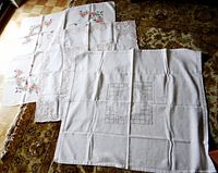 Three tablecloths laid out on floor: hand embroidered floral, machine cutwork, white linen embroidered.
