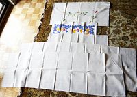 Five table linens laid out: two square embroidered cloths with floral and chick designs, and three runners including a smaller crochet-edged runner and two long matching runners.
