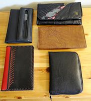 Five leather items laid out on a wooden surface showing two black wallets (one with floral flap), one brown wallet, a red and black wallet, and a zippered black wallet.
