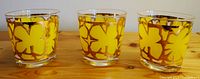 Set of three glasses showing bright yellow floral patterns with gold outlining on clear glass, placed on a wooden surface.