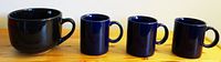 Four ceramic mugs arranged in a row on a wooden surface, showing the size and color difference between one large bluish-black mug and three smaller dark blue mugs.