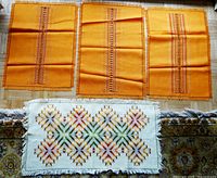 Four table runners displayed on a wooden floor: three orange runners with central vertical stripe detail, one white runner with multicolor diamond pattern and fringe.