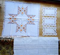 Photo showing all five table linens laid out on floor: the large square embroidered cloth with fringe, the long runner, three smaller lace-edged embroidered mats