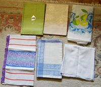 All 5 folded tablecloths and 1 table runner displayed on floor, showing variation in color, pattern, and edging styles.