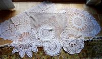 Seven assorted crocheted table mats arranged on floor showing variety of shapes and intricate crochet lacework patterns, including two smaller round ones and one square beige-grey mat.