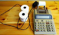 View of Casio calculator connected with charger and showing 2 full paper rolls and one partial roll on wood surface.