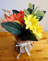 Front view of colorful artificial flower arrangement with bright orange and yellow flowers, green leaves, and small artificial berries in a burlap-wrapped pot tied with a white ribbon.