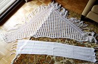 White triangular crochet shawl with lace-like stitching and 6-inch fringe edges, displayed spread on floor.