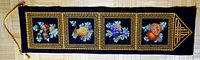 Full view of black rectangular wall hanging with four square framed fruit cross-stitch designs and gold decorative border, with hanging cord visible