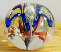 Clear glass paperweight showing vibrant blue swirls with red, white, green, and orange internal decorations on a wooden surface.