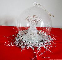 Glass ball ornament with snowflake etching, featuring a glass angel inside. Silver bead garland around base for display.