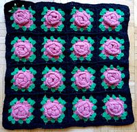 Full view of crocheted pillow cover top with 15 raised pink crocheted flowers grouped in a 3x5 grid on a black crocheted background with green leaves.