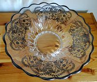 Top down photo of a large clear glass bowl with a scalloped rim and black floral and urn designs near the rim, placed on a wooden surface