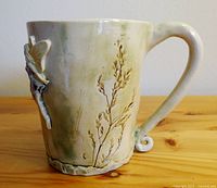 Side view of ceramic mug showing 3D fairy figurine and botanical detailing on off-white surface with slight green patina.