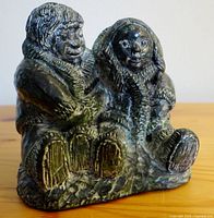 Front-left angled view of the dark stone sculpture showing two Inuit children seated with detailed clothing and facial features.