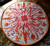 Full view of the round white cotton tablecloth with a symmetrical red and orange floral and leaf design and orange border, spread on a surface.