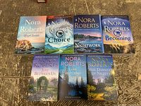 Top front view of all 7 paperback books by Nora Roberts laid out on a wooden floor with covers facing up, showing the titles and cover artwork.