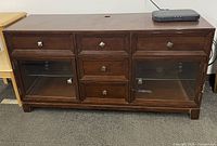 Full view of dark wood media console featuring 5 drawers and 2 glass cabinet doors with metal knobs, rectangular legs, and a cable management hole on the top.