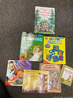 Seven illustrated children's books including educational reading and math books, fairytale, and Christian themed books laid out on carpet