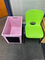 Green children's chair and pink storage box with chalkboard front, placed side by side on carpeted floor near glass window and wicker basket.