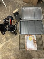 Overview of kitchen utensils in a metal container next to three rectangular metal cooling racks and a box grater with wooden handle on concrete floor.
