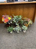 Front-facing view of 6 artificial plants with flowers and foliage in various containers on carpet in front of wooden desk