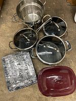 Photo of cookware set including three black Cuisinart pots/pans with glass lids and a stainless steel strainer basket, along with Pampered Chef bakeware items.