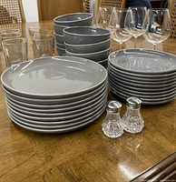 Full view of the dinnerware and glassware set on a wooden table showing stacks of large and small gray plates, bowls, clear drinking glasses, wine glasses, and salt shakers.