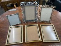 Photo showing six picture frames arranged on wooden table, three gold-tone rectangular frames in front, and three ornate silver-tone frames behind.